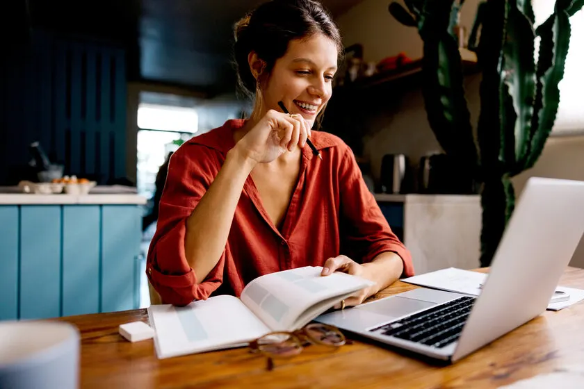 A woman is sitting at a table with an open laptop, writing notes in a notebook.